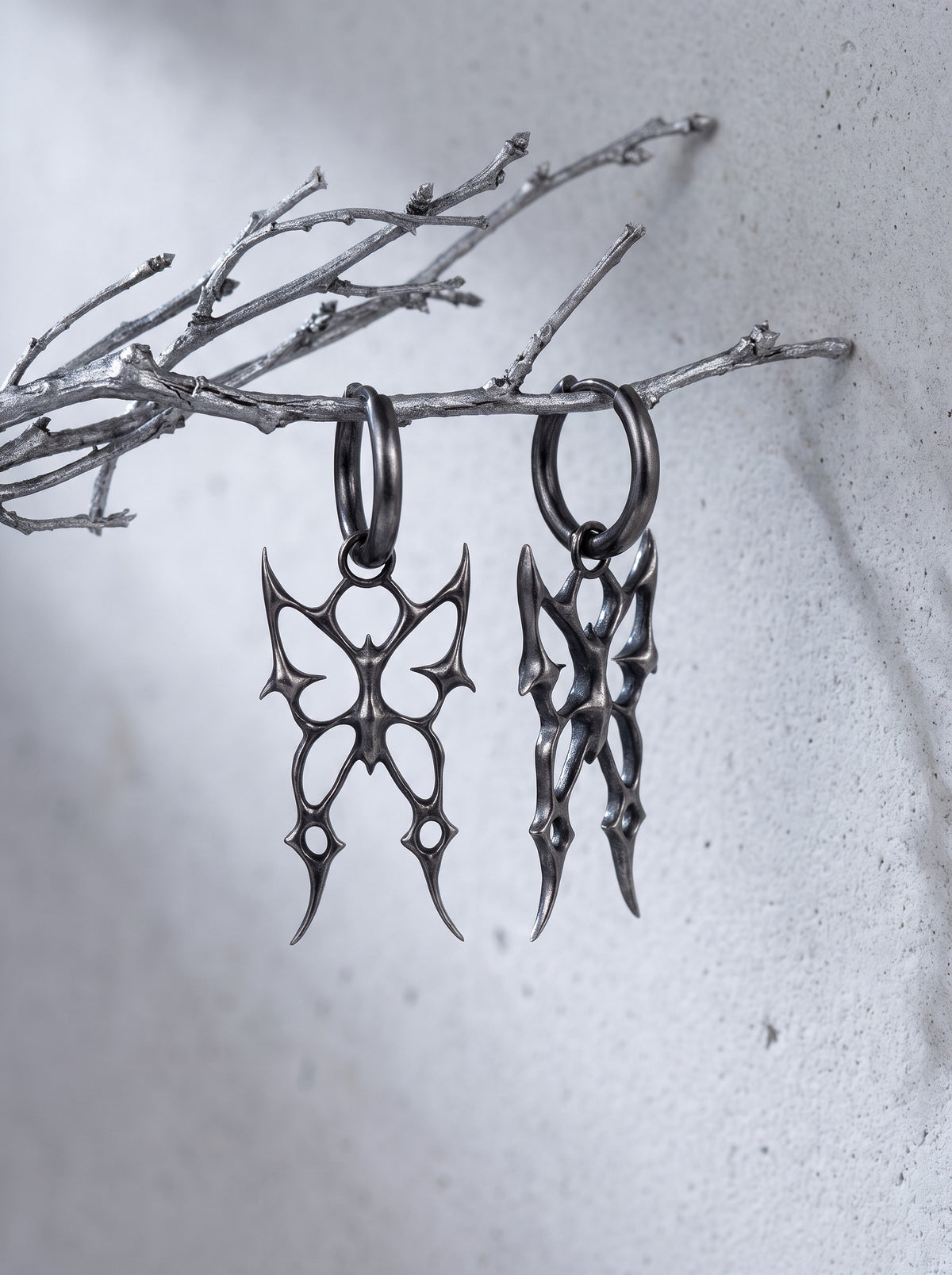 Barbed Butterfly Earrings in oxidised sterling silver hanging from a silver twig against pale concrete