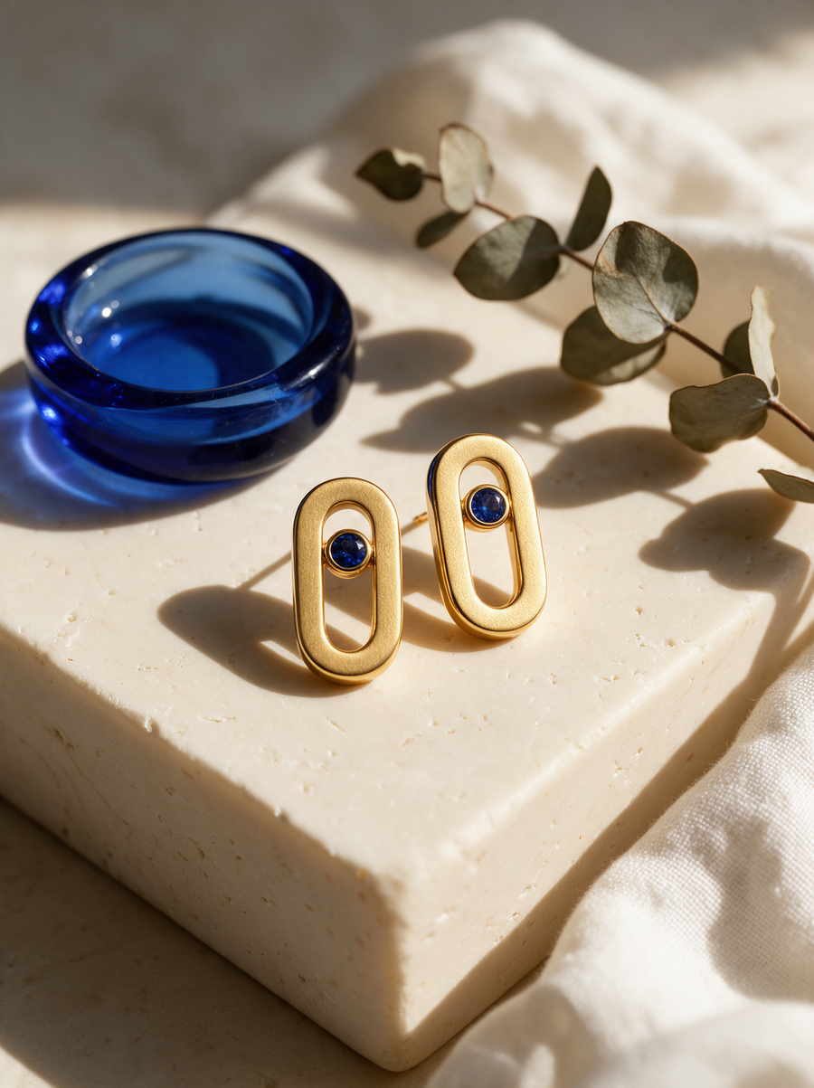 Bolt Studs in 18K gold vermeil with blue stones on a stone block beside a blue glass dish and eucalyptus