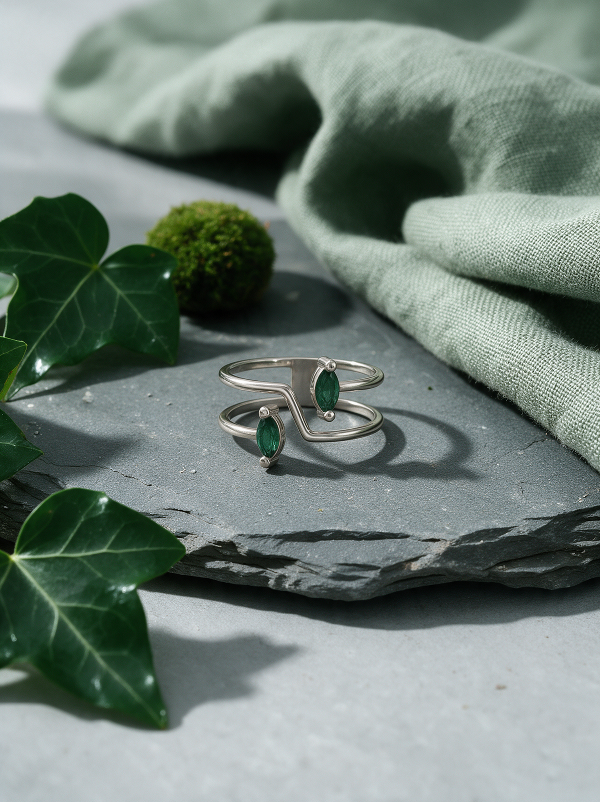 Switchback Ring in sterling silver with green marquise stones on slate beside ivy and sage linen